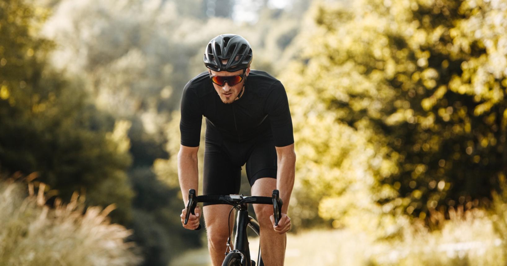 Ciclista su strada in tenuta nera, immerso nella natura, con casco e occhiali.