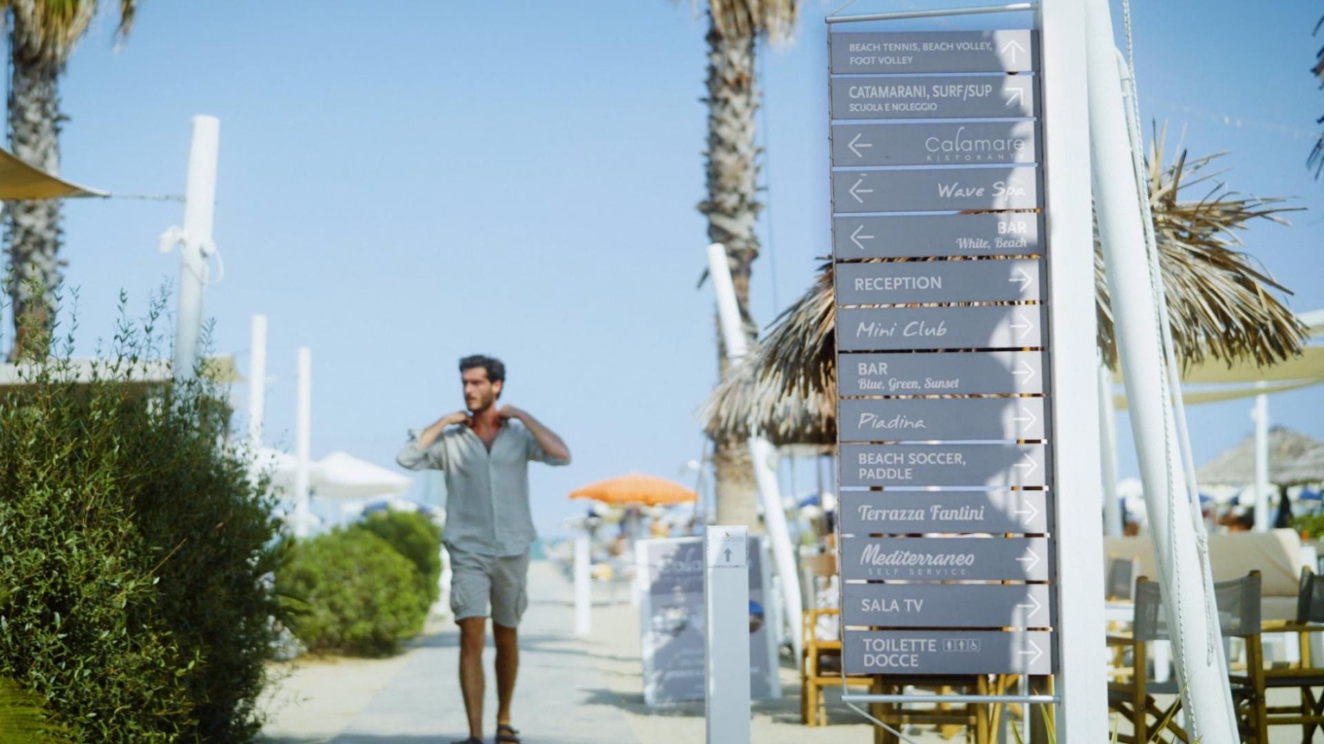 Spiaggia con cartelli direzionali per attività e servizi, uomo che cammina sul sentiero.