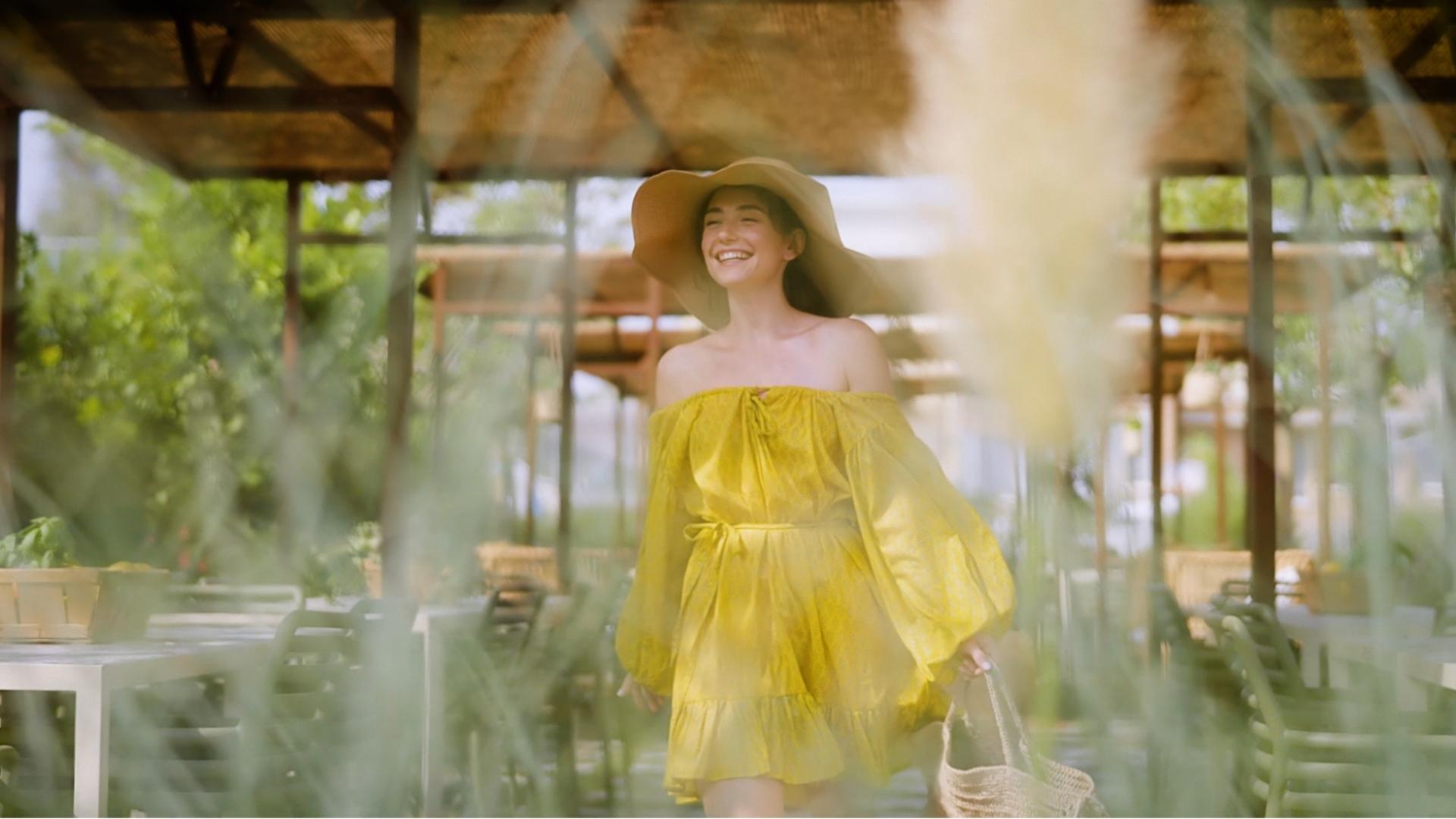 Donna sorridente con abito giallo e cappello, in un ambiente estivo e luminoso.