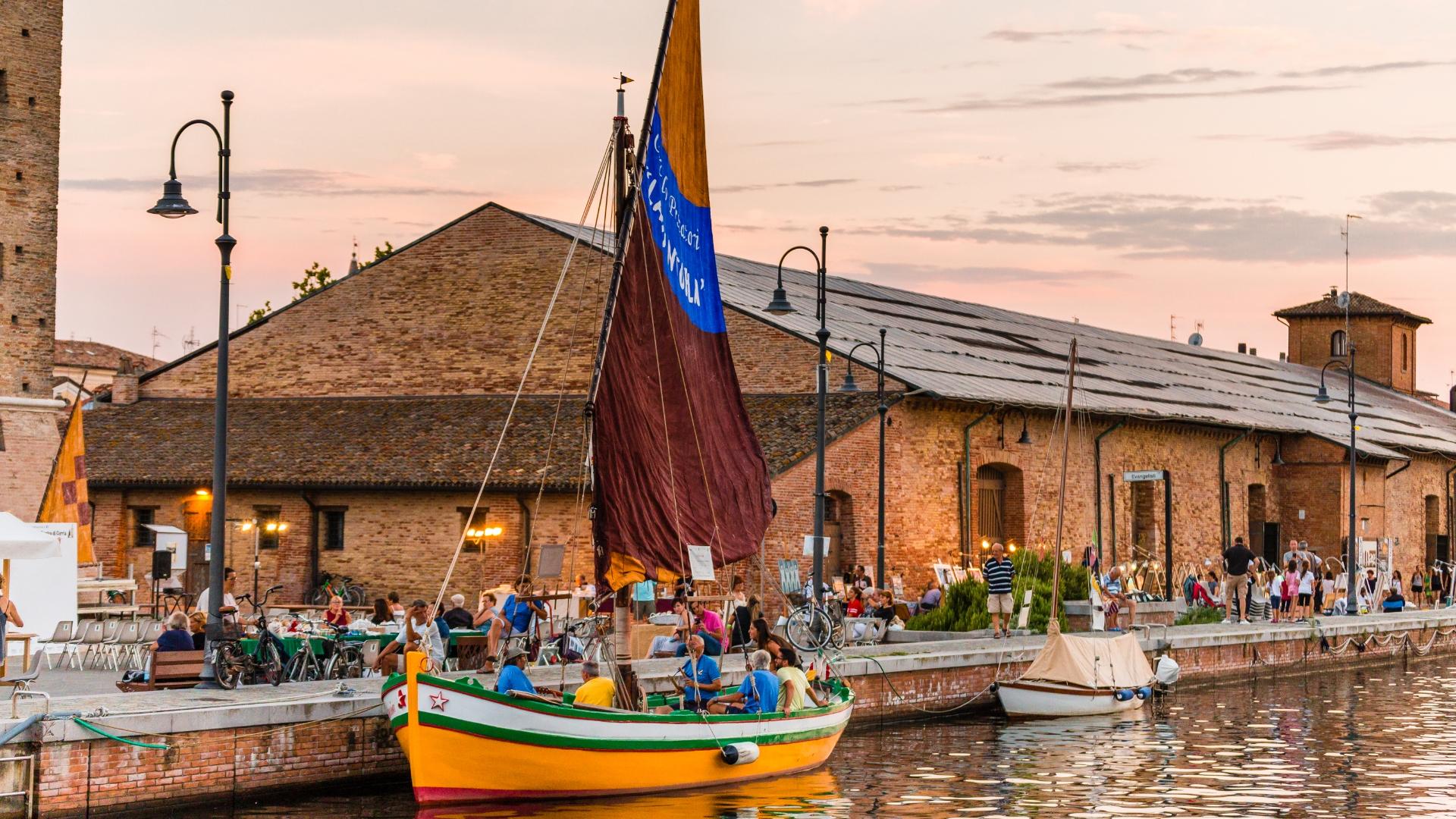 Barca a vela colorata in un porto affollato, al tramonto, vicino a edifici storici.