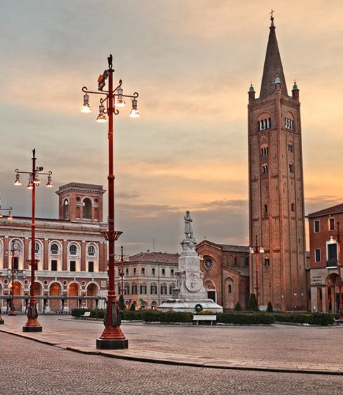 Piazza con edificio storico e torre, illuminata da lampioni al tramonto.