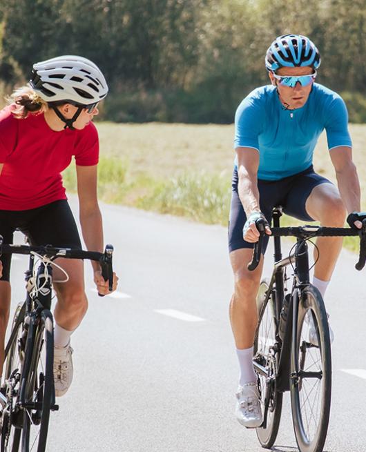 Due ciclisti pedalano su una strada soleggiata, indossando caschi e abbigliamento sportivo.