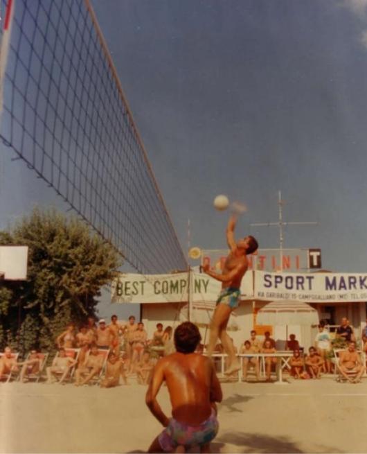 Partita di beach volley estiva con pubblico e insegna di un negozio sullo sfondo.