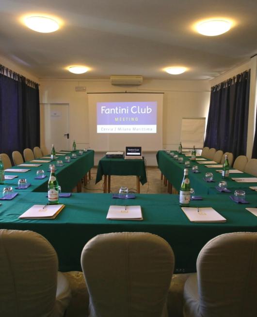 Sala conferenze con tavoli verdi, blocchi appunti e proiettore, pronta per un meeting.