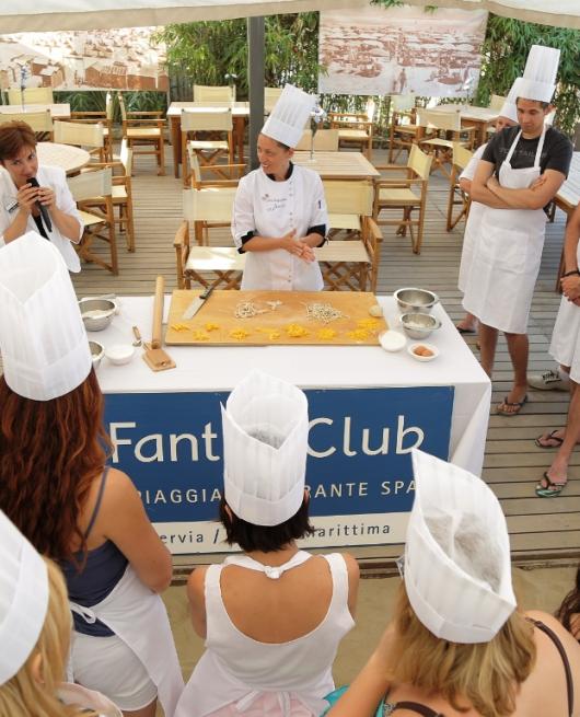 Lezione di cucina con chef e partecipanti in grembiule e cappello da cuoco.