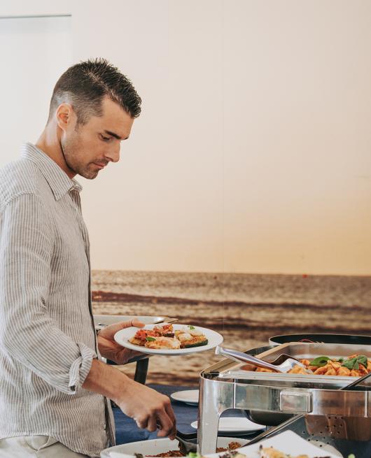 Uomo serve cibo da un buffet con piatti vari e vista mare sullo sfondo.