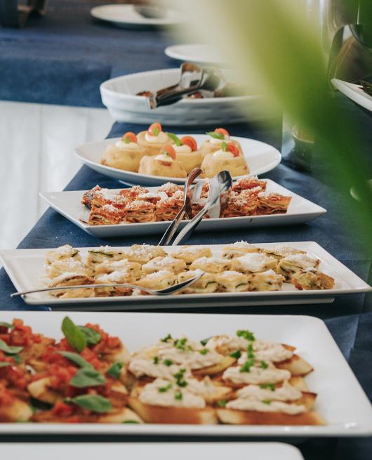 Buffet di antipasti con bruschette, lasagne e verdure grigliate su piatti bianchi.