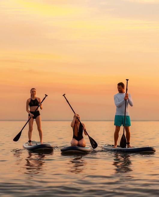 Tre persone fanno paddleboarding al tramonto su un mare calmo.