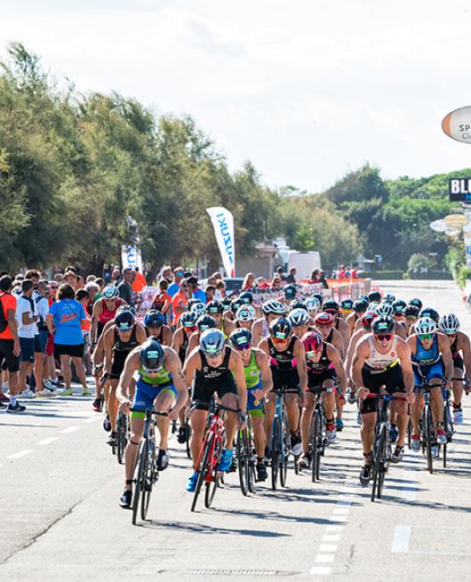 Gara ciclistica con molti partecipanti e spettatori lungo la strada alberata.