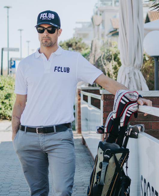 Uomo con cappello e polo FCLUB, occhiali da sole, accanto a una sacca da golf.