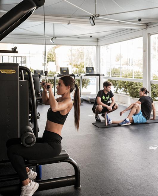 Persone si allenano in palestra con pesi e attrezzi, ambiente luminoso e spazioso.