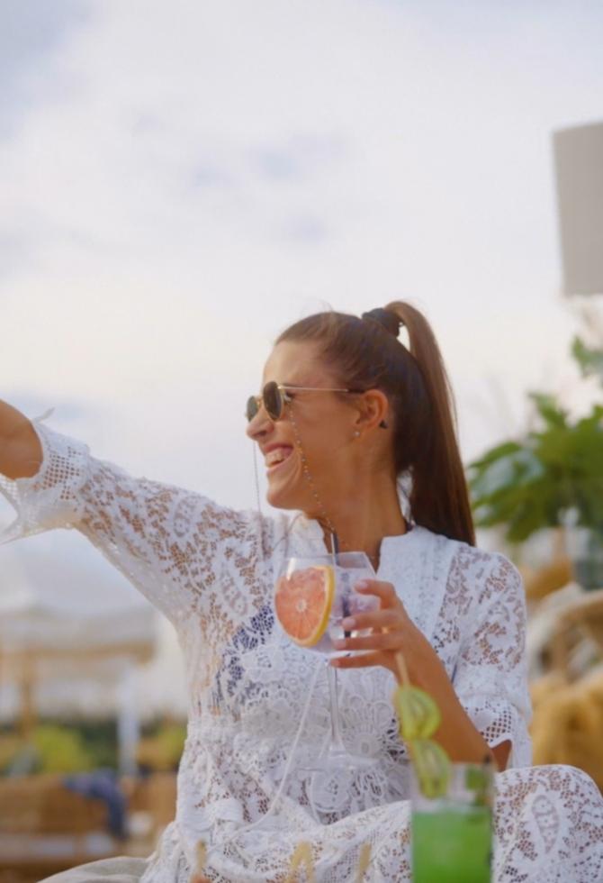 Donna sorridente in abito bianco, con occhiali da sole e drink fruttato in mano.